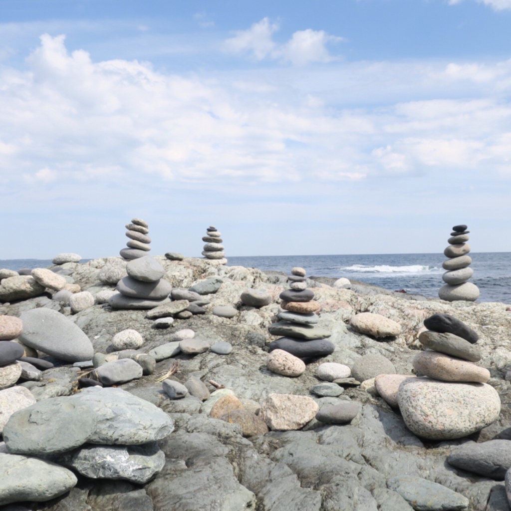 Several stacks of "zen tower" stones can be seen on top of a large rock. Some of them are intact and some appear to have fallen over, scattering their stones around. In the background, the ocean and the sky are visible. 