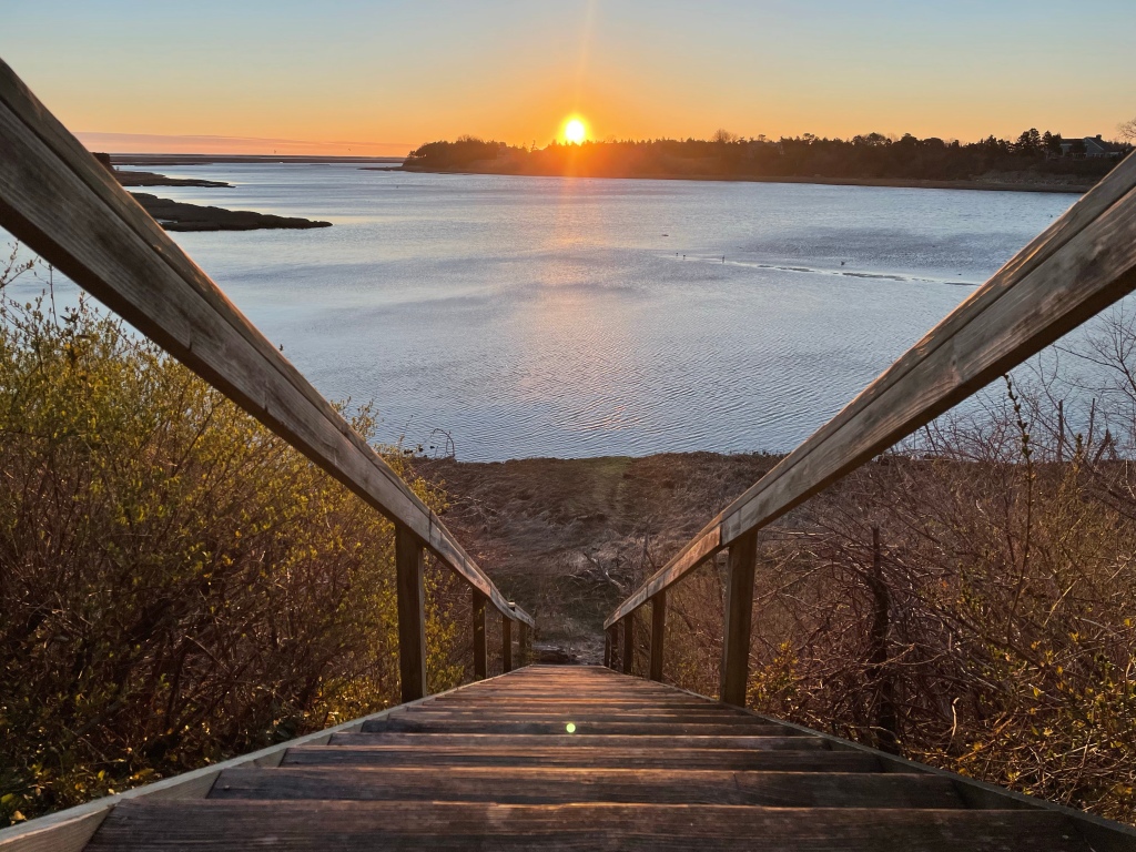 This is an image of wooden stairs going down to a marshy beach area. There are bushes to the sides of the stairs. The water is dark blue-gray with many small ripples. There are several dark inlets of land visible in the bay. The sun is orange, rising over the middle of the far side of the scene. 