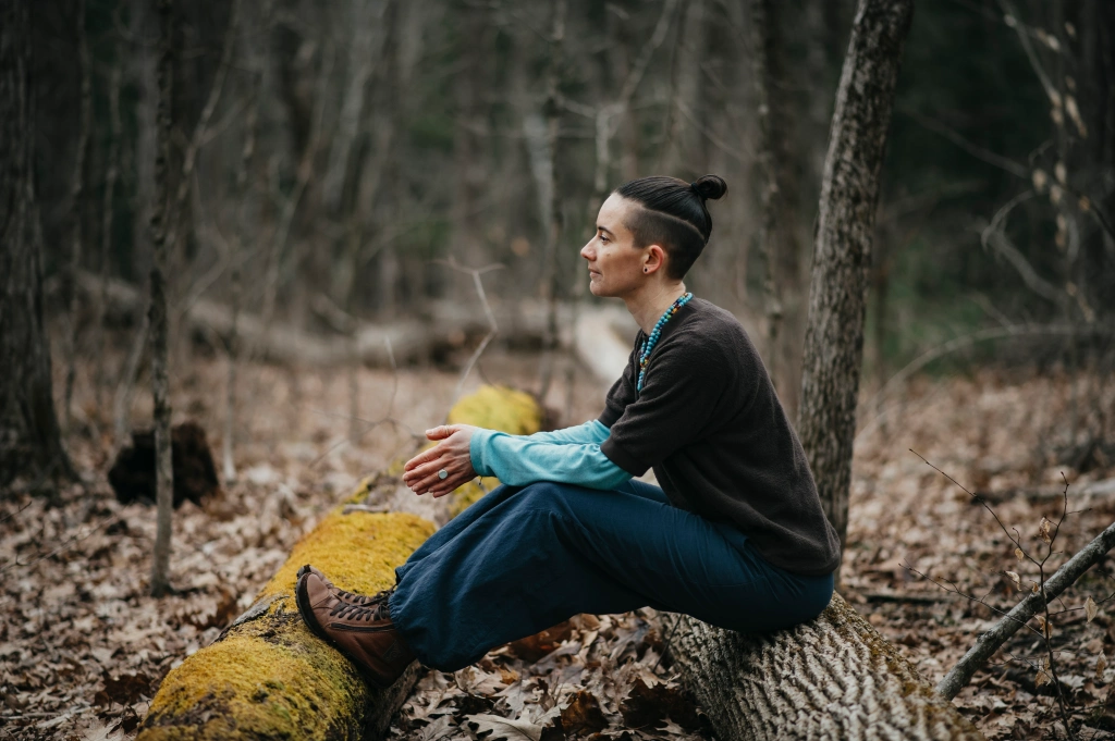 This is an image of the same nonbinary person as the header of the home page. This image is in color. The person is sitting on one log with their feet up on another log. The log with their feet on it is covered in bright green-yellow moss. They are looking to the side so their left side is visible. Their sneakers are brown leather, pants dark blue, long sleeve dark brown shirt over turquoise long sleeve shirt. Their beads are blue. An aquamarine ring is visible on their left ring finger. Their hair is pulled back in a bun, cut in an undercut. They have a neutral face expression. Behind them are blurry trees and twigs in the woods. 