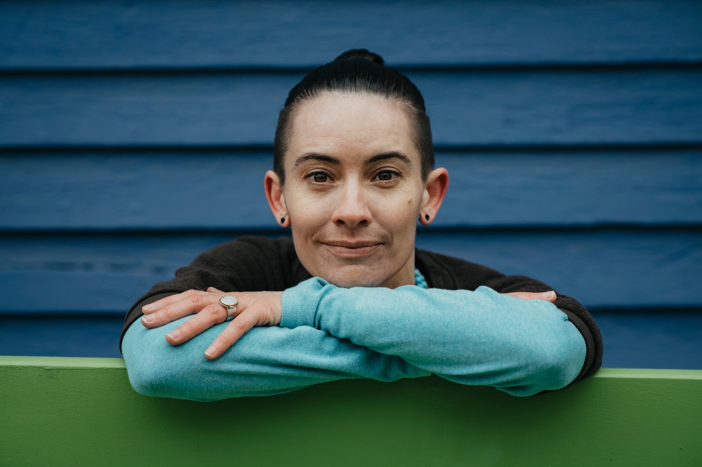 A nonbinary person with dark brown hair pulled back into a bun, dark brown eyes, and lightish color skin is facing directly into the camera. They have a slight smile on their face. They are leaning down on their crossed arms which rest on the back of a green bench. They have a brown short sleeve shirt on over a turquoise long sleeve shirt. There is a blue siding wall behind them. An aquamarine ring is visible on their left hand.