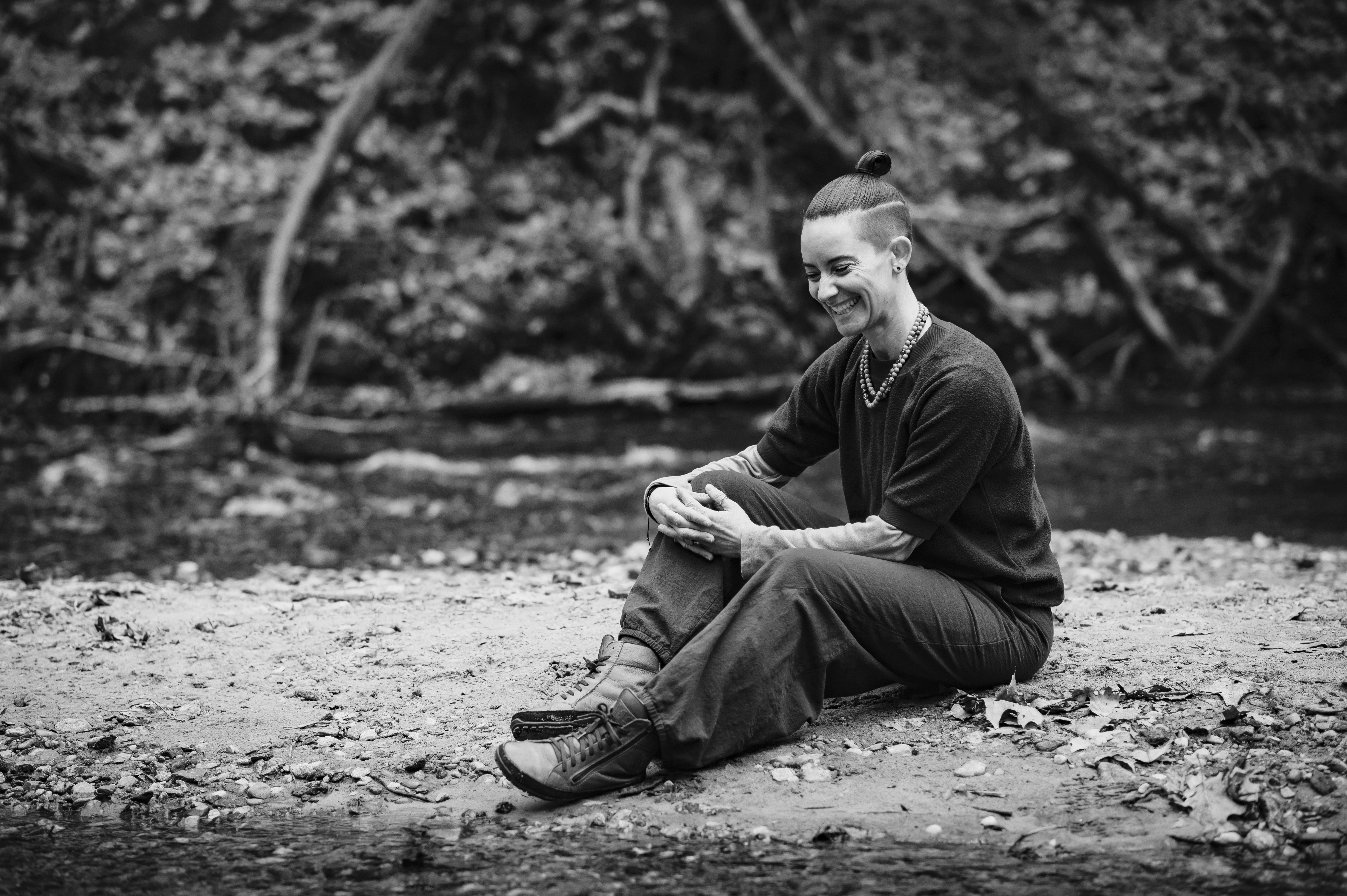 This is a black & white grayscale photograph of a nonbinary person. The person is seated on the ground, facing slightly angled to their right so their left side is visible. The person is smiling, communicating happiness and warmth. They are looking towards the ground which is dirt with stones and leaves. Their hair is an undercut with the top part pulled back into a bun. They have a beaded necklace on. They are wearing a darker colored short sleeve shirt over a lighter colored long sleeve shirt. There is a stream and the opposite bank with tree roots in the background.